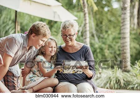 Picture - Older Caucasian woman and grandchildren looking at photo album. Fotosearch