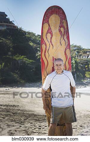 Stock Photo - Older Hispanic man with surfboard on beach. Fotosearch