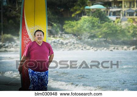 Stock Photo - Older Hispanic man with surfboard on beach. Fotosearch