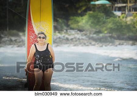 Stock Photo - Older Hispanic woman with surfboard on beach. Fotosearch