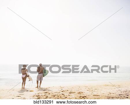 Stock Image - Couple carrying surfboards on beach. Fotosearch