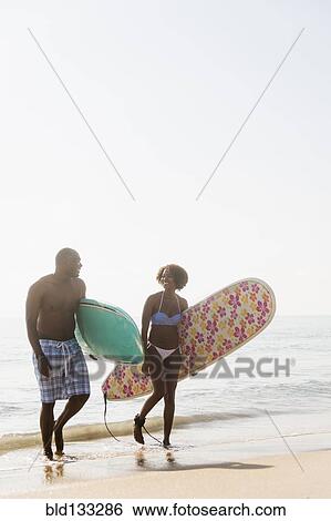 Stock Photograph - Couple carrying surfboards on beach. Fotosearch