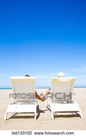 Stock Image - Couple holding hands in lounge chairs on beach. Fotosearch