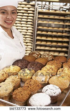 Stock Image - Hispanic baker carrying tray of muffins in bakery. Fotosearch