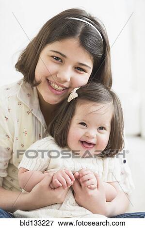 Stock Photo - Hispanic girl holding toddler sister in living room. Fotosearch
