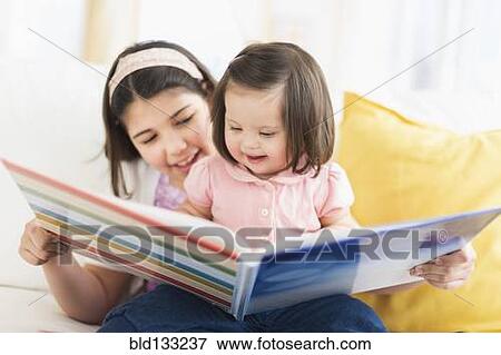 Stock Photo - Hispanic girl reading to toddler sister in living room. Fotosearch