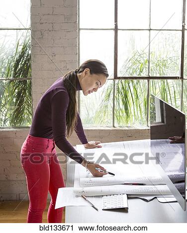 Stock Image - Mixed race architect examining blueprints in office. Fotosearch