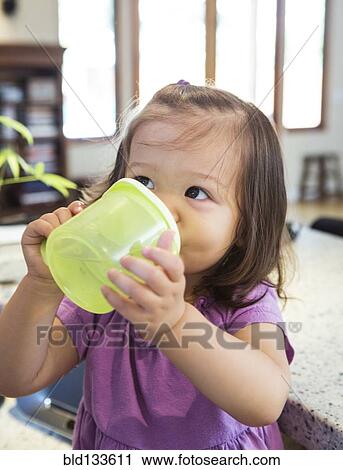 Stock Image - Mixed race girl drinking from sippy cup in kitchen. Fotosearch