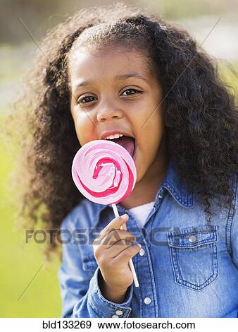 Stock Photo - Mixed race girl licking lollipop outdoors. Fotosearch