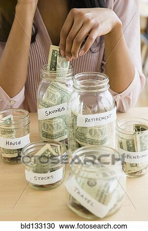 Stock Image - Mixed race woman putting money in savings jars. Fotosearch