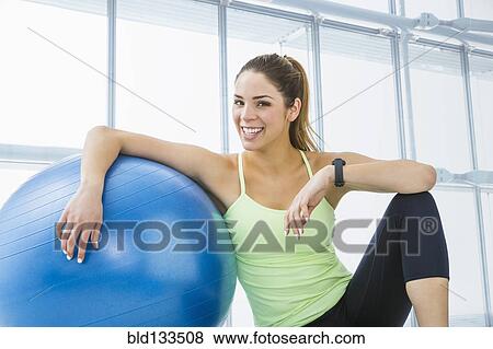 Stock Photo - Mixed race woman resting with exercise ball in gym. Fotosearch