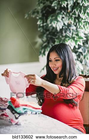 Stock Image - Pregnant Asian woman picking out baby clothes. Fotosearch