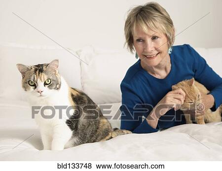 Stock Photo - Senior Caucasian woman petting cats on bed. Fotosearch