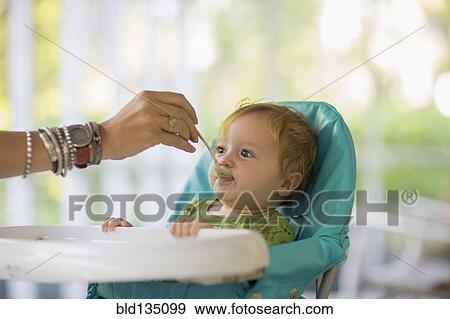 Stock Photo - Caucasian mother feeding baby in high chair. Fotosearch