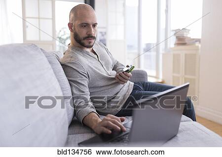 Stock Photograph - Hispanic man using cell phone and laptop on sofa. Fotosearch