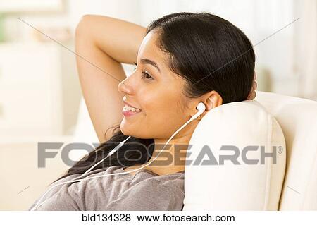 Stock Photo - Hispanic woman listening to earphones on sofa. Fotosearch