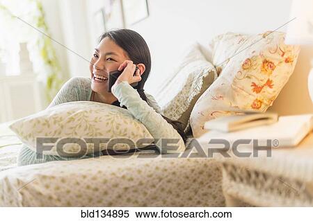 Stock Photography - Mixed race teenage girl talking on cell phone on bed. Fotosearch