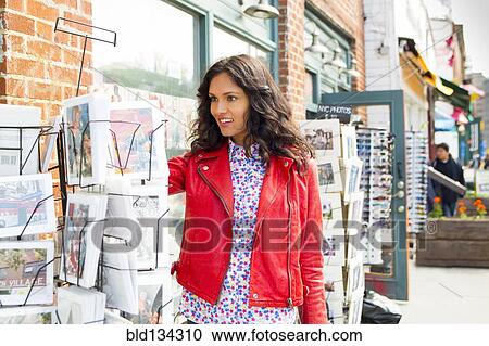 Stock Image - Mixed race woman browsing postcards on city sidewalk. Fotosearch