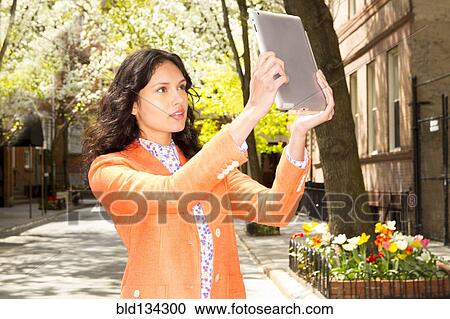 Stock Image - Mixed race woman taking picture with tablet computer on city street. Fotosearch