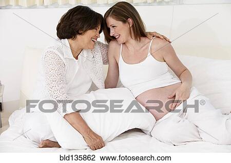 Stock Image - Mother hugging pregnant daughter on bed. Fotosearch