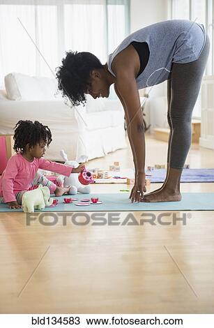 Stock Image - Mother stretching as daughter plays on yoga mat. Fotosearch