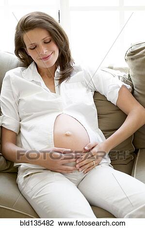 Stock Image - Pregnant woman holding her belly on sofa. Fotosearch