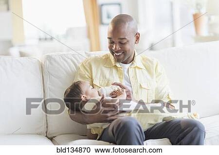 Stock Image - Smiling father feeding baby on sofa. Fotosearch