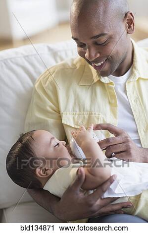 Stock Image - Smiling father feeding baby on sofa. Fotosearch