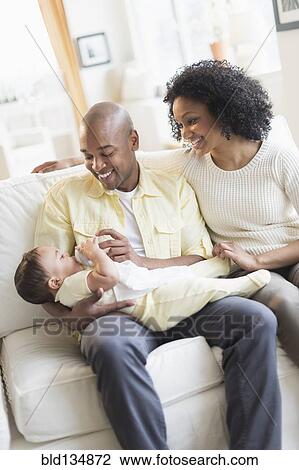 Stock Image - Smiling father feeding baby on sofa. Fotosearch