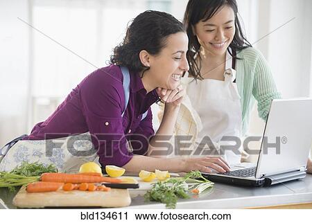 Stock Image - Women using laptop and cooking together. Fotosearch