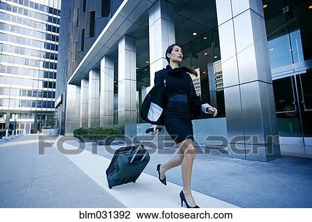 Stock Image - Mixed race businesswoman rolling baggage in city. Fotosearch