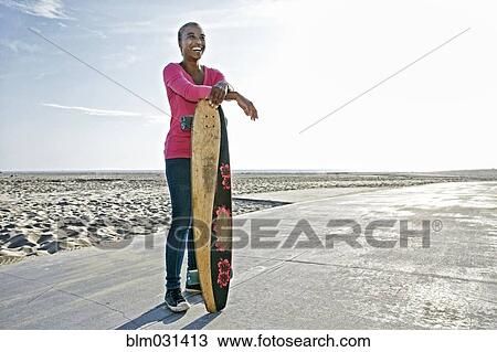 Stock Image - Older Black woman holding skateboard on beach. Fotosearch