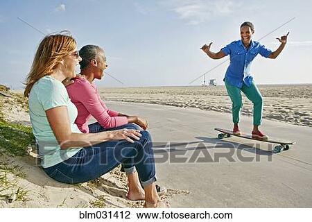 Stock Image - Older Black woman skateboarding by friends on beach. Fotosearch
