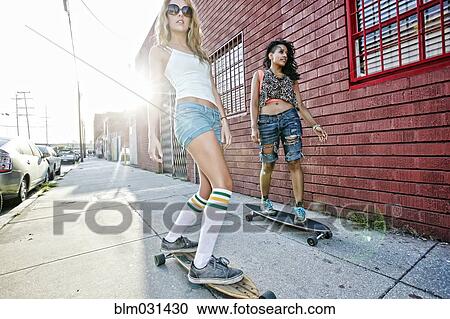 Stock Image - Women riding skateboards on city street. Fotosearch