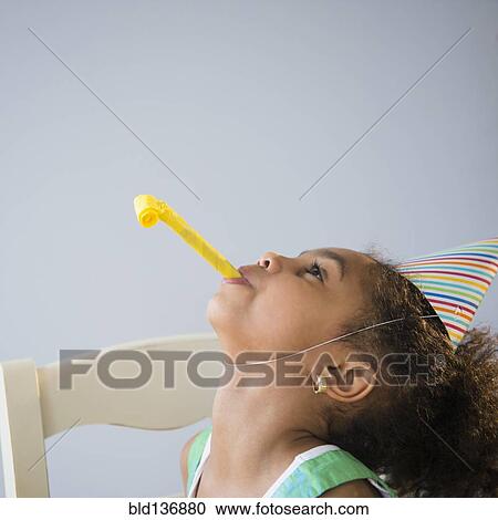 Stock Image - African American girl blowing noisemaker at birthday party. Fotosearch