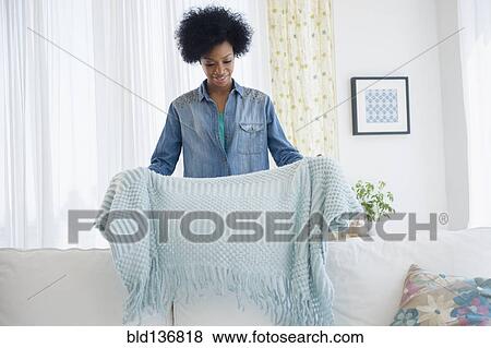 Stock Photo - African American woman folding blanket on sofa. Fotosearch