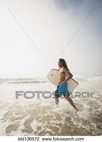 Stock Image - Black teenage boy carrying boogie board in waves. Fotosearch