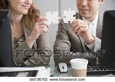 Stock Image - Business people putting puzzle pieces together at desk. Fotosearch