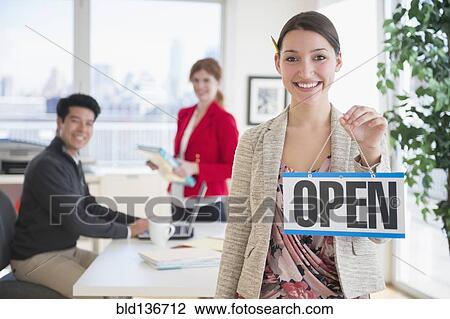 Stock Image - Businesswoman holding Open sign in office. Fotosearch