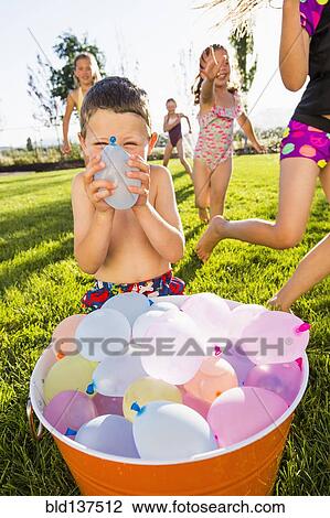 Stock Image - Caucasian children playing with water balloons in backyard. Fotosearch