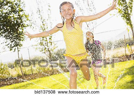 Stock Photo - Caucasian girls playing in sprinkler in backyard. Fotosearch