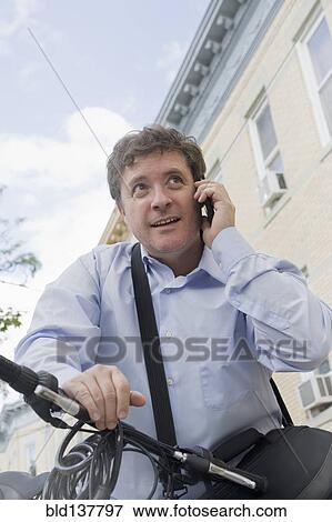 Stock Photo - Hispanic businessman talking on cell phone on bicycle on city street. Fotosearch