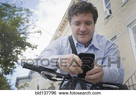 Stock Photograph - Hispanic businessman using cell phone on bicycle on city street. Fotosearch