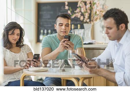 Stock Image - Hispanic friends using cell phones in cafe. Fotosearch