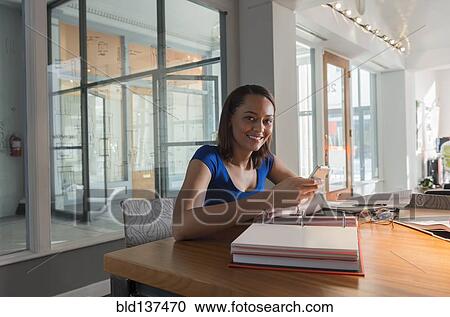 Stock Image - Mixed race businesswoman using cell phone at desk. Fotosearch