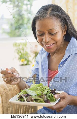 Stock Image - Mixed race woman eating salad on sofa. Fotosearch