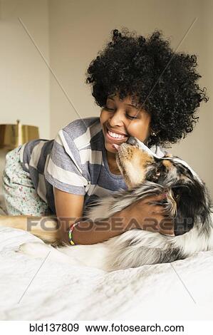 Stock Photo - Mixed race woman playing with dog on bed. Fotosearch
