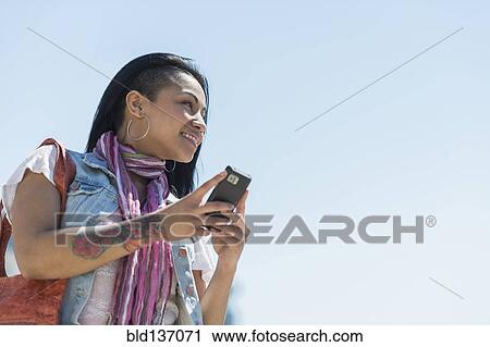 Stock Image - Mixed race woman using cell phone under blue sky. Fotosearch