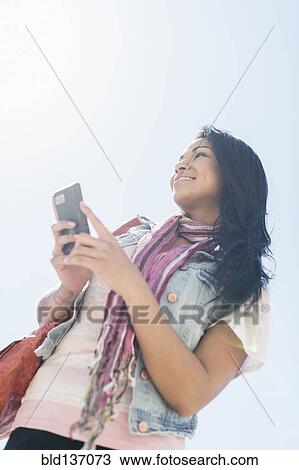 Stock Image - Mixed race woman using cell phone under blue sky. Fotosearch