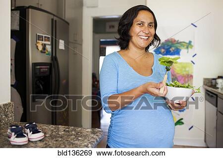 Stock Photo - Pregnant Hispanic woman eating salad in kitchen. Fotosearch